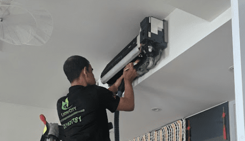 The photo shows a technician cleaning the indoor unit of a split-type air conditioner, using professional tools to remove dirt and buildup while performing maintenance.